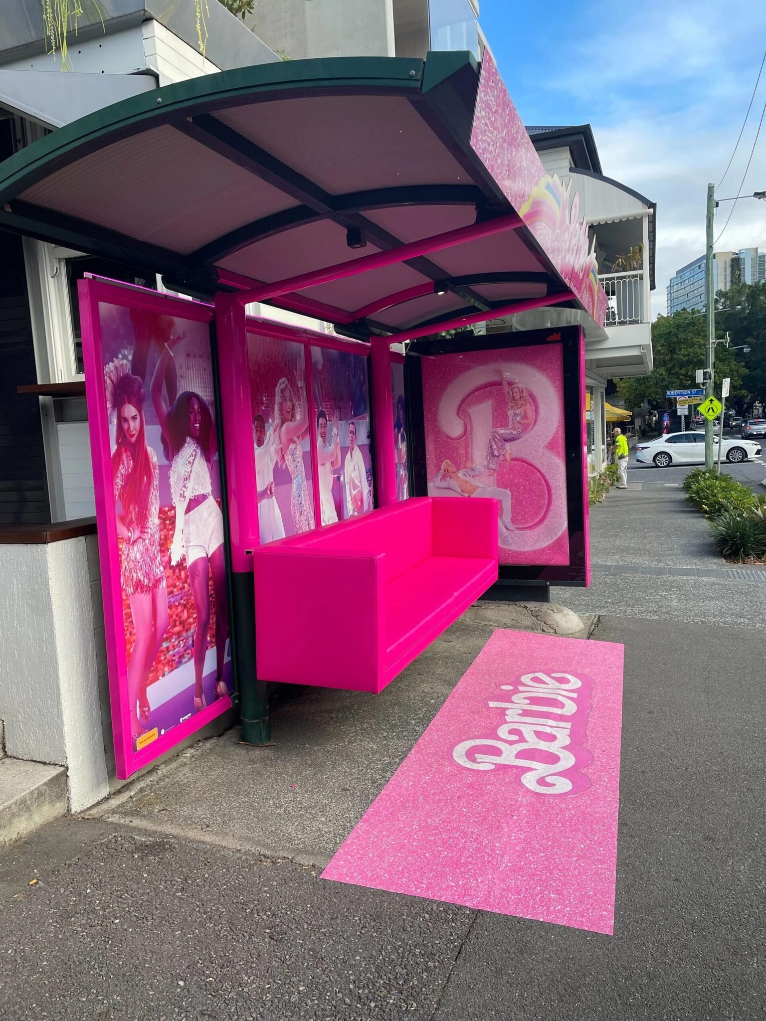 Front of a Barbie-themed storefront with bright pink seating, large fashion posters, and a pink Barbie carpet on the sidewalk.