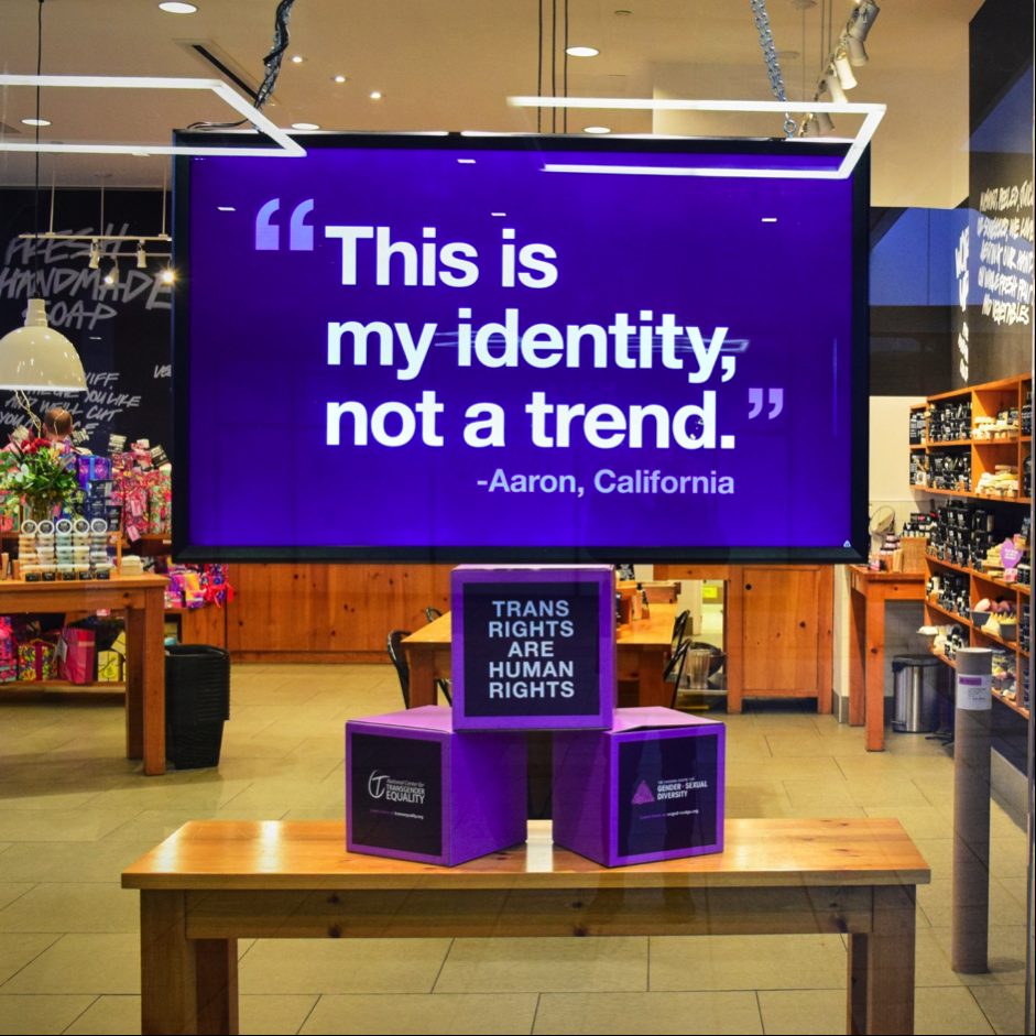 Large purple sign in a store reads: 'This is my identity, not a trend.' – Aaron, California, with purple boxes below declaring 'Trans rights are human rights.' Social marketing campaign leaning into activism. 