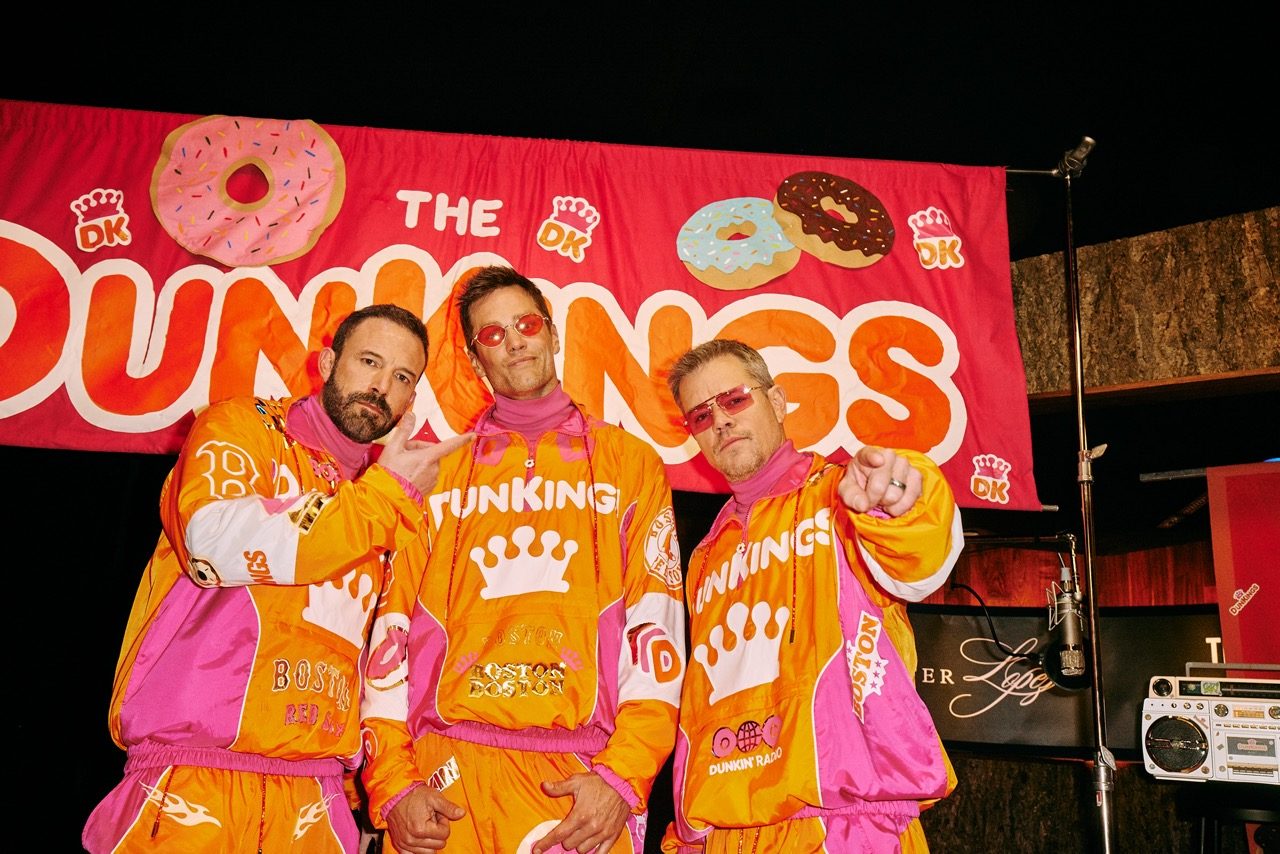 Three men in bright orange-pink Dunkin' outfits pose on a stage in front of a large pink donut banner with the word 'DUNKINGS' visible as a social marketing campaign.