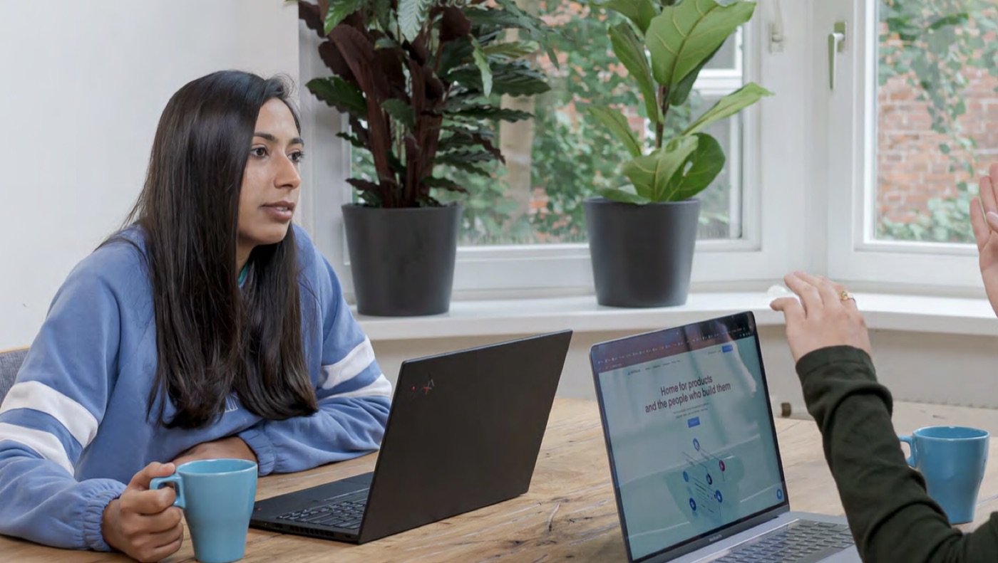 Two colleagues talking across a table, each with a laptop and a blue mug