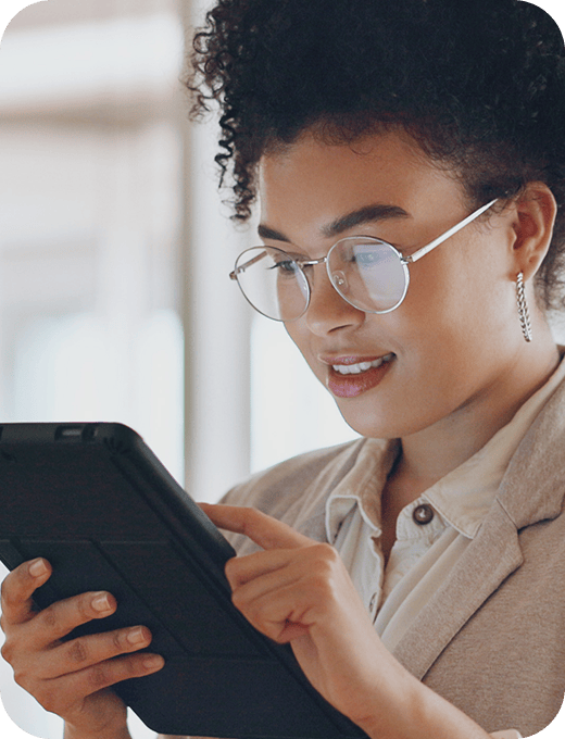 A young woman wearing glasses and business casual attire taps on a tablet screen in a well-lit office setting.